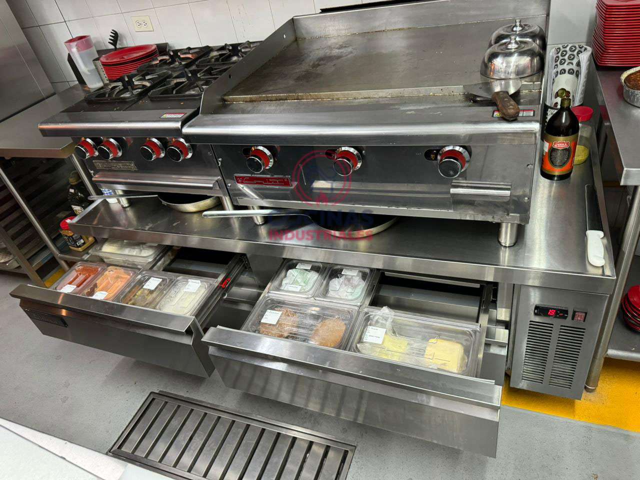 Una configuración de cocina comercial cuenta con una estufa de acero inoxidable con múltiples quemadores y una plancha. Debajo de la estufa, hay cajones del refrigerador abiertos que muestran varios alimentos almacenados en recipientes transparentes. Encima de la estufa se colocan cuencos rojos y una tetera.