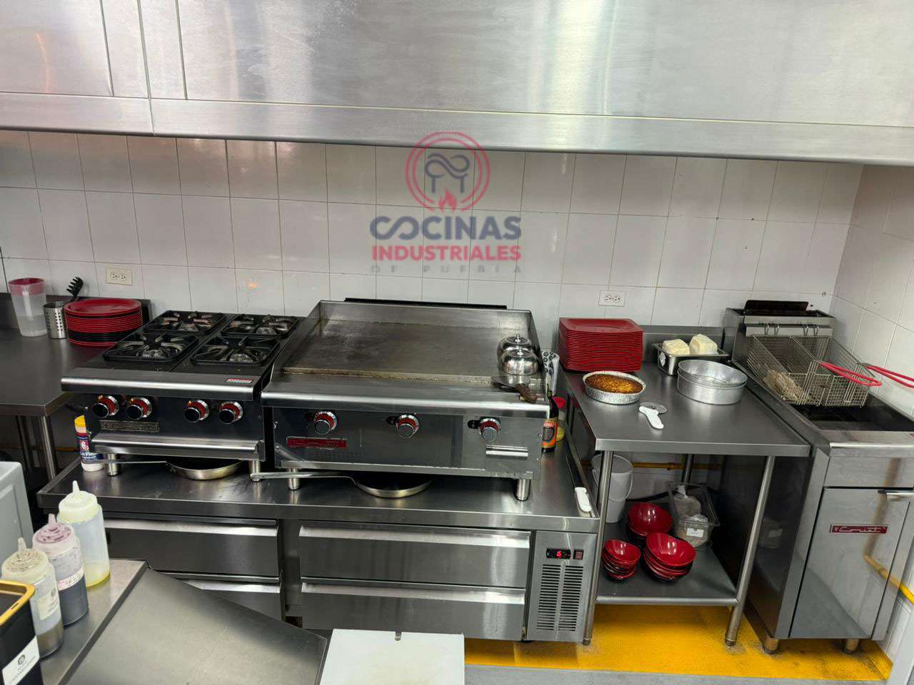 Una cocina comercial con estufa, plancha, freidora y varios utensilios de cocina. Ollas, sartenes y recipientes están cuidadosamente dispuestos sobre las encimeras y estantes. Hay un logo de "Cocinas Industriales" en la pared al fondo.