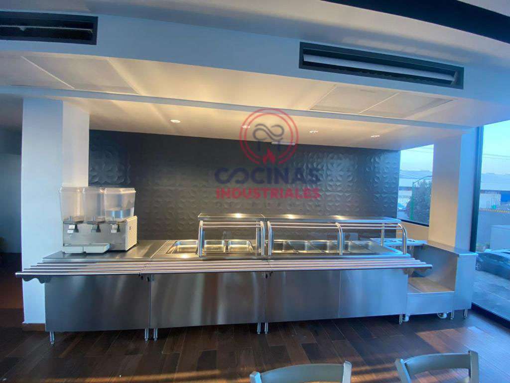 Una configuración de cocina industrial moderna con una encimera de acero inoxidable con varias bandejas para calentar alimentos y un dispensador de bebidas a la izquierda. La cocina tiene una elegante pared con dibujos negros y está iluminada por luces empotradas. El área del bar tiene las palabras "Cocinas Industriales" y un logo encima.