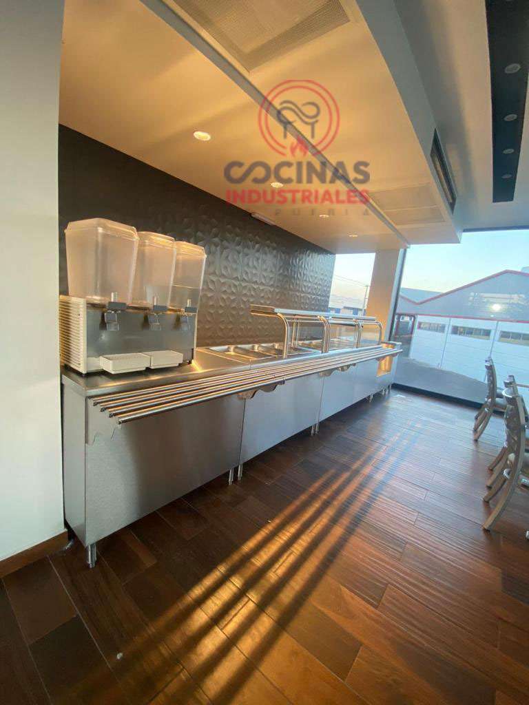 Un mostrador de servicio de alimentos de acero inoxidable está equipado con dispensadores de bebidas y almacenamiento debajo de los estantes. El mostrador está ubicado en una sala moderna con pisos de madera y grandes ventanales, lo que permite que la luz natural ilumine el espacio. El logo de "Cocinas Industriales" es visible arriba.
