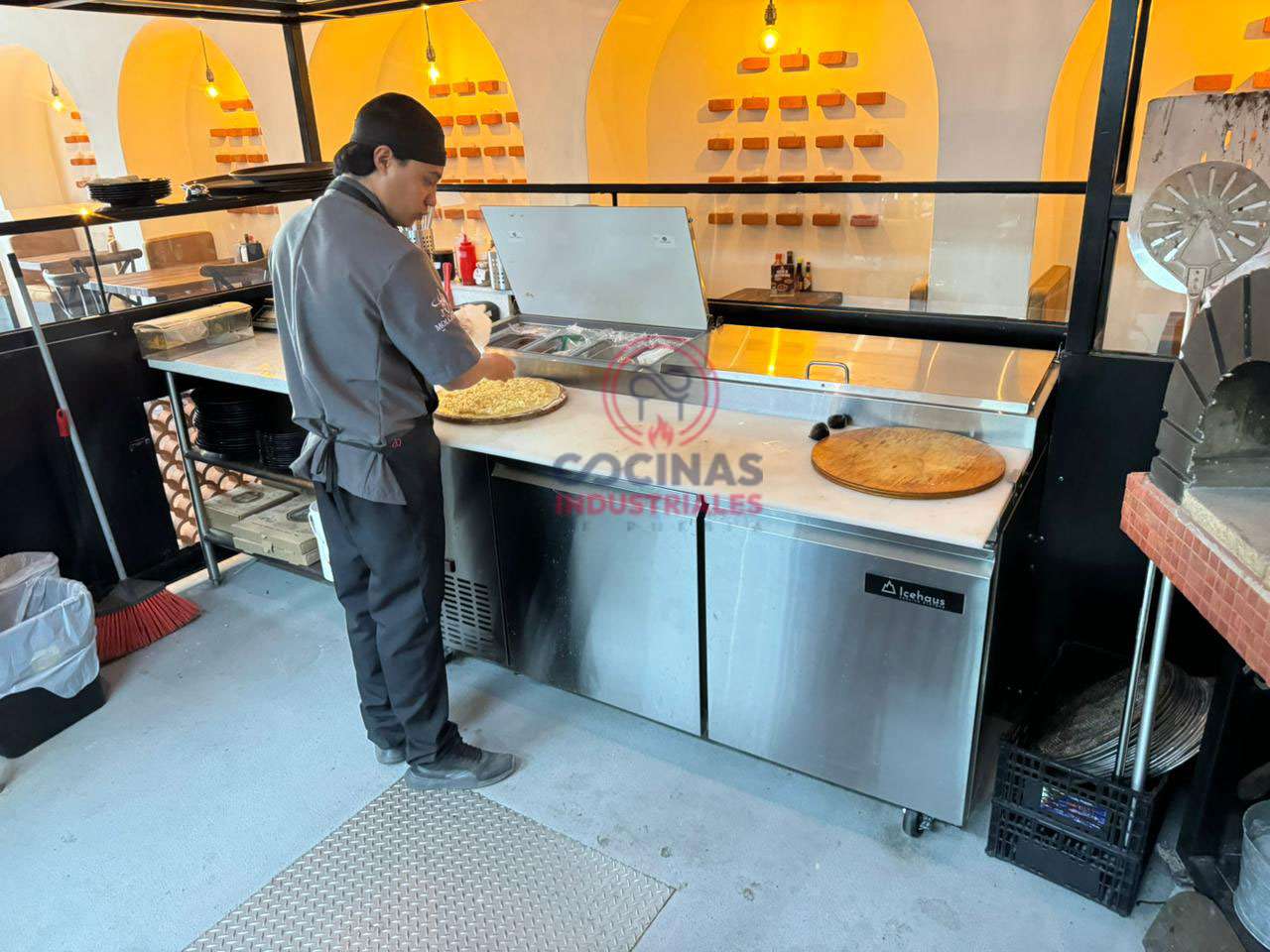 Un chef prepara comida sobre una encimera de acero inoxidable en una cocina comercial. El escenario incluye varios ingredientes y utensilios, un horno de pizza a la derecha y un especiero cuidadosamente dispuesto al fondo. El chef viste un sombrero negro y un uniforme oscuro.